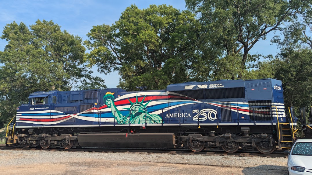 A Norfolk-Southern train engine painted blue with red, white, and pale blue striping behind a painting of the Statue of Liberty and the slogan, America 250.