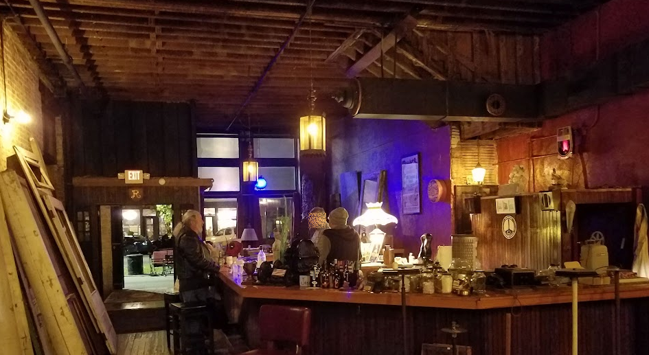 The entryway to a bar, taken from the rear, showing three men standing around an fishook-shaped bar. 