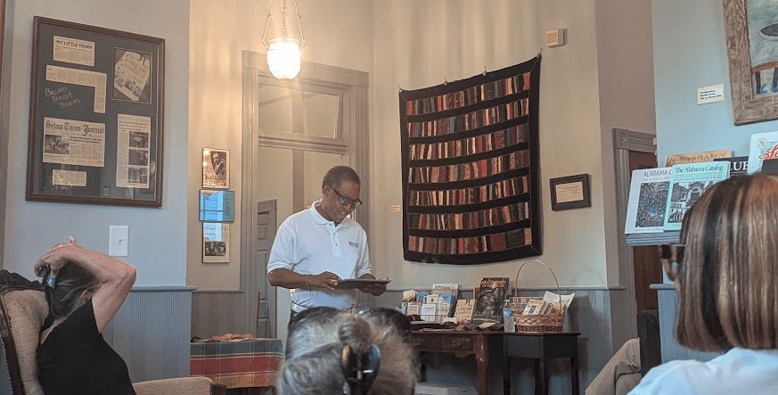 A man reading from a book with a book-themed quilt behind him