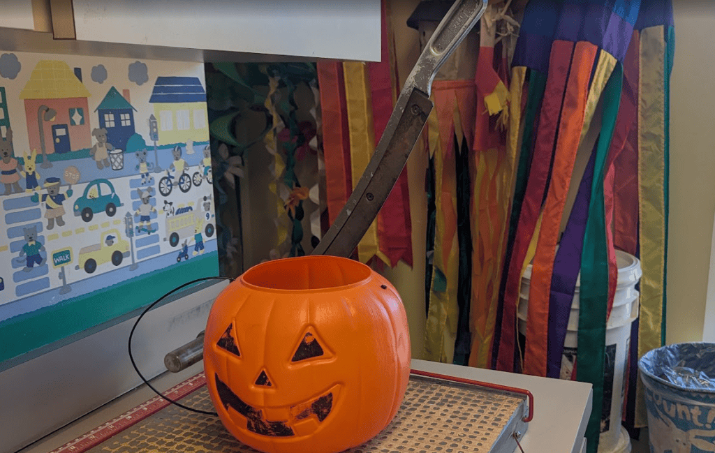 A plastic Jack o Lantern bucket sitting on an old paper cutter with a stout fixed blade behind it
